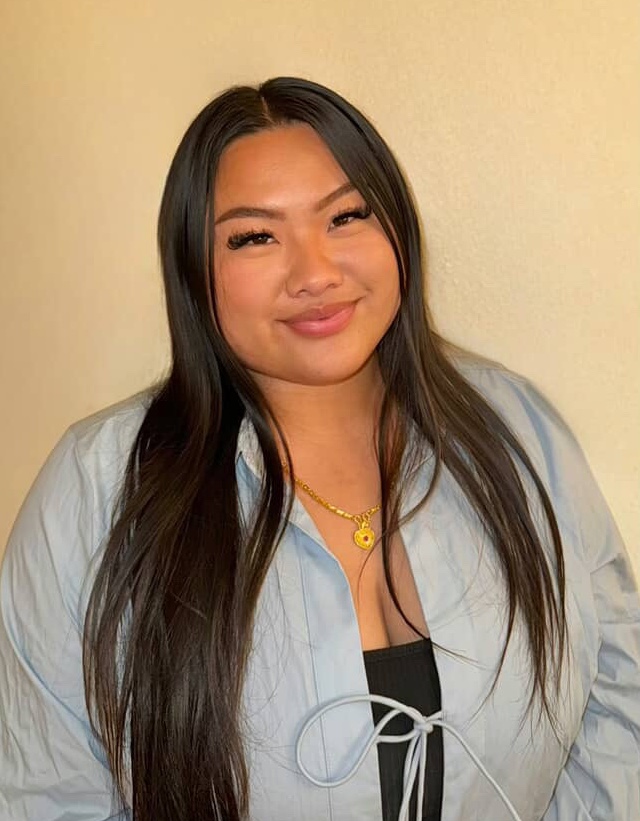 This is a headshot of alumna, Kathy, who wears a blue blouse. Kathy's hair is down and she stands against a plain wall.