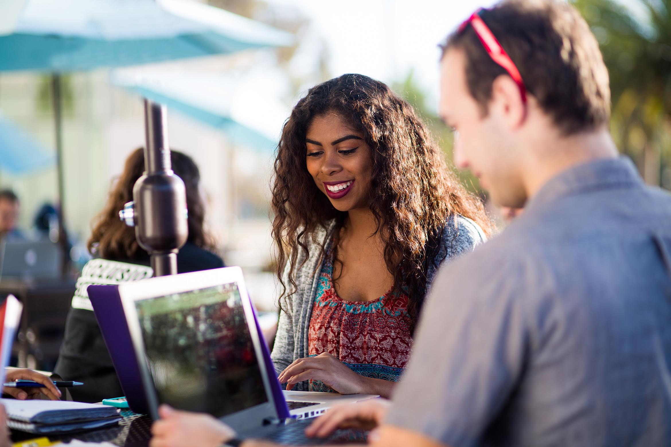 Two students smile while working outdoors on their laptops.