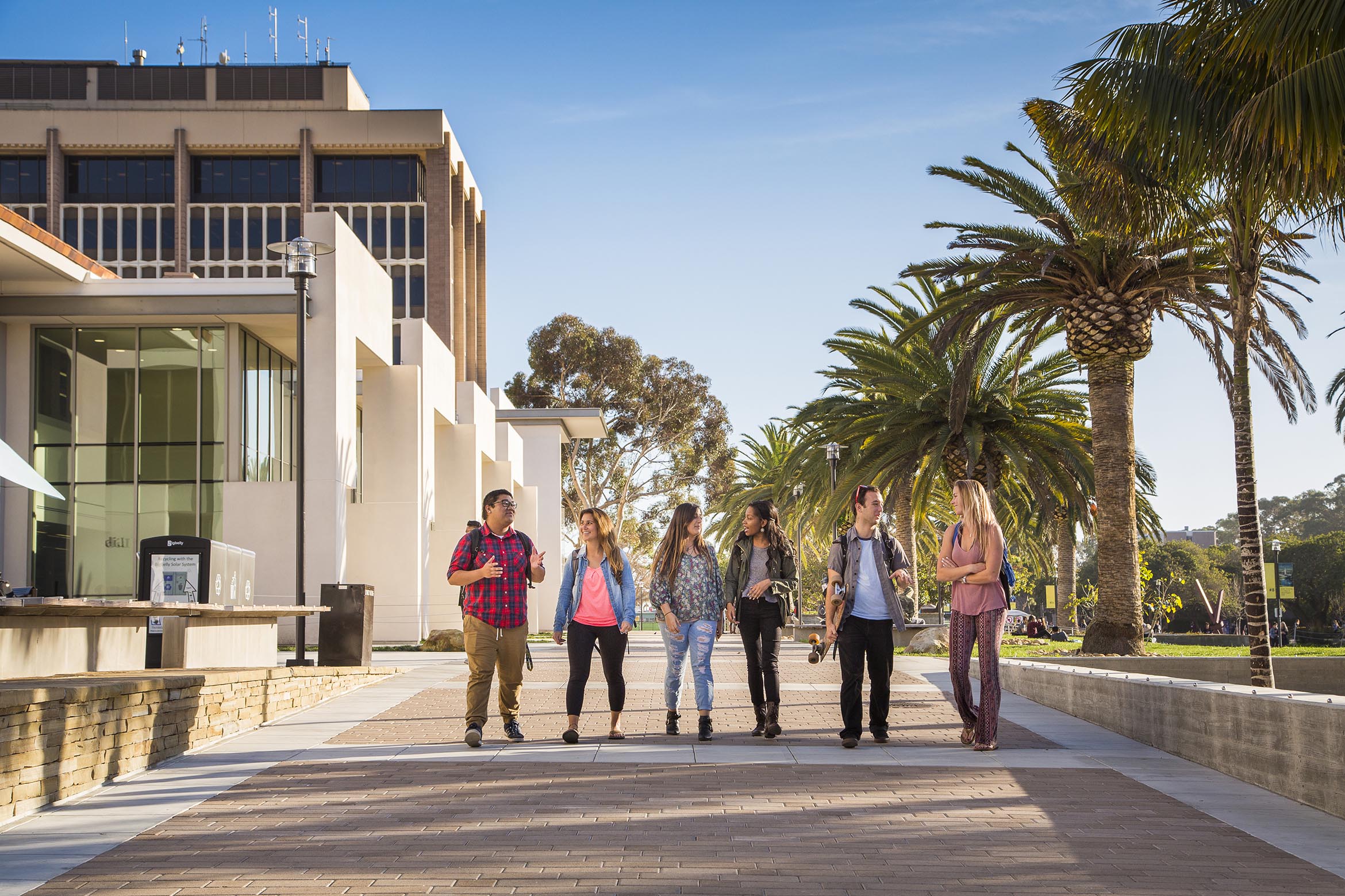 Six students in pairs converse as they walk in front of the library.